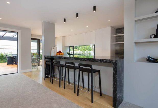 Open-plan contemporary kitchen and living area with a long dark stone island, white full-height cabinetry, timber flooring, and four black bar stools. Recessed and pendant-style ceiling lighting, open shelving, and large glazing connect the kitchen to an outdoor terrace. The space is bright, clean, and refined with strong black-and-white contrast softened by warm timber.