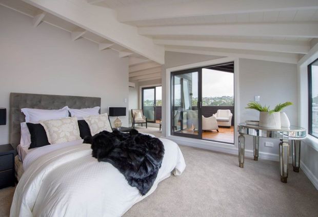 Bright, spacious contemporary bedroom with a large upholstered bed, layered white bedding, patterned pillows, and a dark faux-fur throw. Exposed white ceiling beams and structural detailing add character, while extensive black-framed glazing opens toward a small balcony or terrace and a distant urban view. A mirrored console with decorative vases sits beside the windows, and a sitting area is visible beyond the glazed partition.