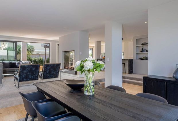 Open-plan contemporary dining and living area with a long dark timber dining table and black upholstered chairs in the foreground. The space features pale walls, a light timber floor, a recessed fireplace, built-in shelving, square structural columns, and large glazed openings connecting to a landscaped outdoor area. Neutral finishes and abundant daylight create a clean, refined atmosphere.
