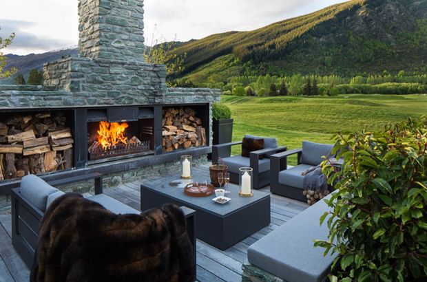 A sophisticated outdoor entertaining area set against a dramatic mountainous landscape. The terrace features a large stacked-stone outdoor fireplace with integrated firewood storage, contemporary grey upholstered lounge seating, and a dark coffee table arranged on grey decking or paving. Lush planting frames the foreground, creating a refined yet natural alpine setting.