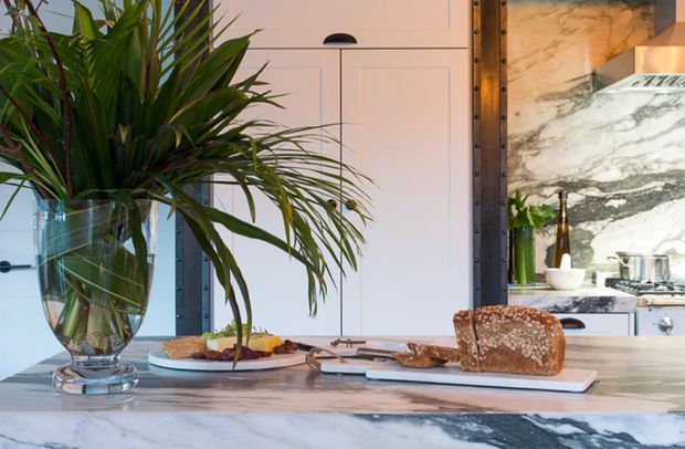 A styled kitchen island scene with a large leafy plant in a glass vase, a marble island benchtop, bread and plated food, and white shaker-style cabinetry. A dramatic veined marble splashback and stainless-steel cooking area are visible in the background, creating a refined, warm contemporary kitchen setting.