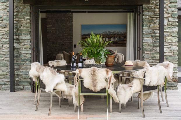 A sophisticated dining area opening onto a stone-clad exterior or terrace. A large round dining table with a reflective dark top is surrounded by upholstered chairs draped in light fur throws. A central leafy plant, tableware, and wine bottles create a warm entertaining setting. The opening is framed in dark timber or metal, with rugged layered stone walls and a light timber deck or floor. A landscape artwork and additional seating are visible beyond.