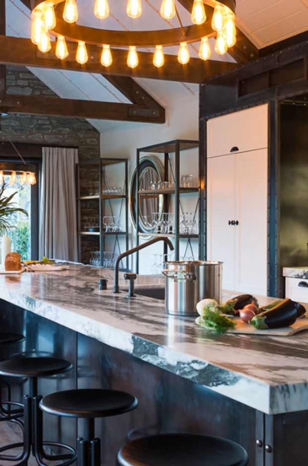 A dramatic, industrial-style kitchen with a large marble island, dark metal cabinetry, black bar stools, open glass shelving, exposed timber beams, and a ring-shaped pendant light with multiple warm bulbs. Stone walls, white ceiling panels, and a tall integrated white cabinet add contrast, while vegetables and cookware sit on the island.
