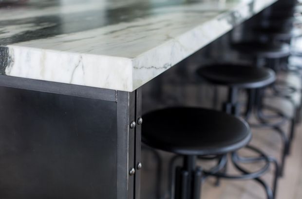 Close-up of a dark, industrial-style kitchen or bar counter with a thick white veined stone benchtop, dark metal base, exposed fasteners, and black round bar stools.