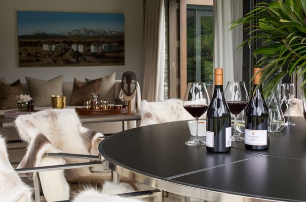 Elegant dining and sitting area with a large round dark dining table set with wine bottles and glasses. White upholstered chairs are draped with plush fur throws, while a lounge area, large landscape artwork, sheer curtains, and abundant indoor greenery create a warm, refined atmosphere.