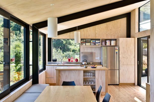 Bright, compact open-plan kitchen and dining area with extensive floor-to-ceiling glazing, pale plywood wall and ceiling linings, integrated cabinetry, a stainless-steel refrigerator, and a timber dining table with built-in window seating. Black structural framing creates strong contrast, while views of greenery surround the space.