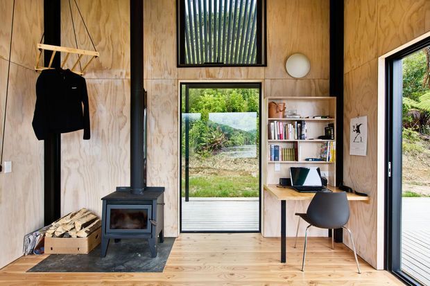 A compact timber-lined multifunctional interior with a small black wood-burning stove, firewood storage, a wall-mounted clothes rail, and a built-in home office nook. Pale plywood wall panels and timber flooring create a warm cabin-like atmosphere. Large black-framed windows and a glazed door open views toward lush vegetation and an exterior deck, while a vertical window with exterior slats adds daylight and privacy.