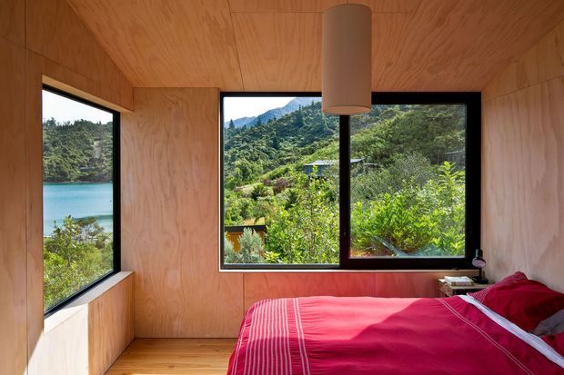A compact bedroom lined in warm plywood, with a low bed dressed in deep red and pink bedding. Large black-framed windows wrap around two walls, bringing in abundant natural light and views of a turquoise body of water, dense vegetation, and surrounding hills. A simple cylindrical pendant hangs centrally from the plywood ceiling, creating a warm, minimalist cabin-like atmosphere.