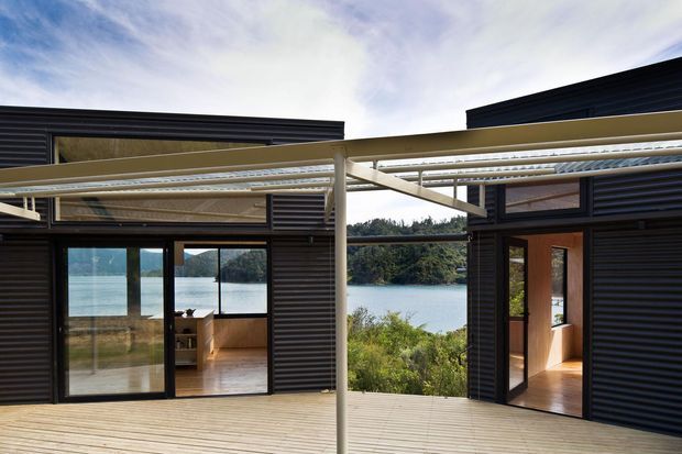 A contemporary dark-clad waterfront house opening onto a broad timber deck. Large sliding glass doors reveal warm timber interiors, while a pale structural pergola or covered walkway spans the foreground. Water and wooded shoreline are visible beyond the house.