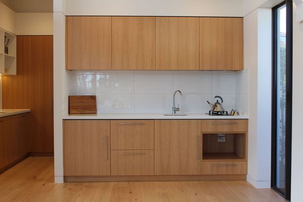 Compact contemporary kitchen with warm timber cabinetry, white tiled splashback, stainless-steel sink and tapware, and timber flooring. Full-height glazing at the right brings in natural light.