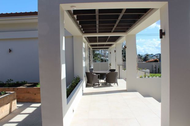A bright, modern covered outdoor terrace or patio viewed through a large rectangular opening. The space has white rendered walls, pale tiled flooring, a dark exposed grid ceiling beneath a pergola-like roof, built-in planter beds, and a small outdoor dining setting. A suburban landscape is visible beyond the terrace.