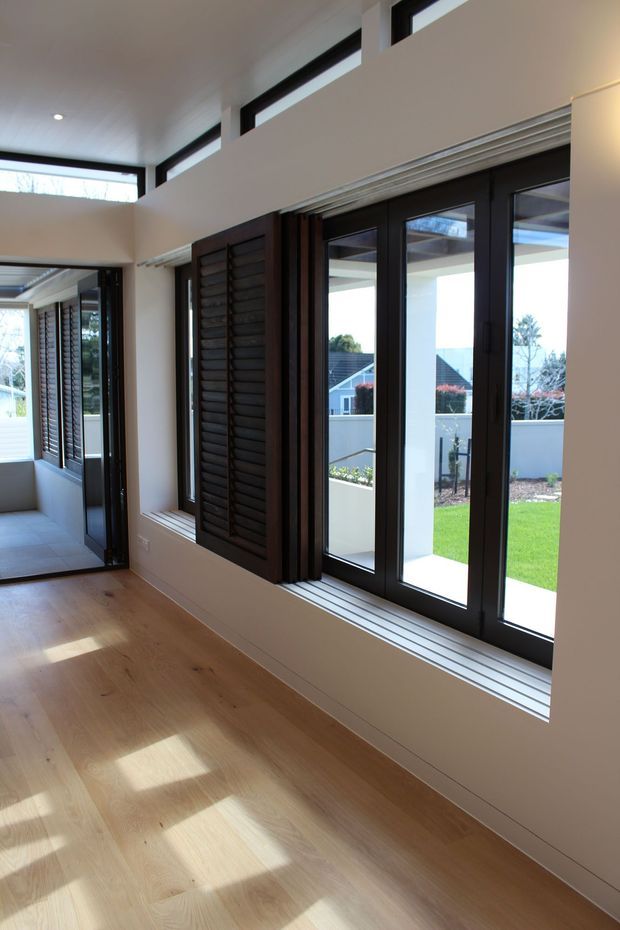 Bright modern interior corridor or living space with pale timber flooring, cream walls, high clerestory windows, and large black-framed glazed doors and windows. Dark timber louvred shutters and sliding or folding glazing line the wall, opening toward a landscaped garden and neighbouring homes. Strong natural daylight creates reflected rectangular patches across the floor.