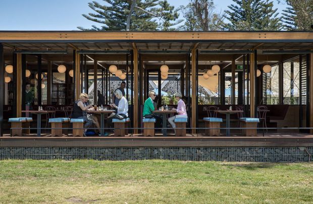 Exterior view of a contemporary open-air restaurant or café pavilion, with extensive glazed folding doors, timber-framed structure, a shallow flat roof, outdoor dining tables and striped bench seating. Warm spherical pendant lights and patterned screens are visible inside, with landscaped lawn and mature trees in the foreground and background.