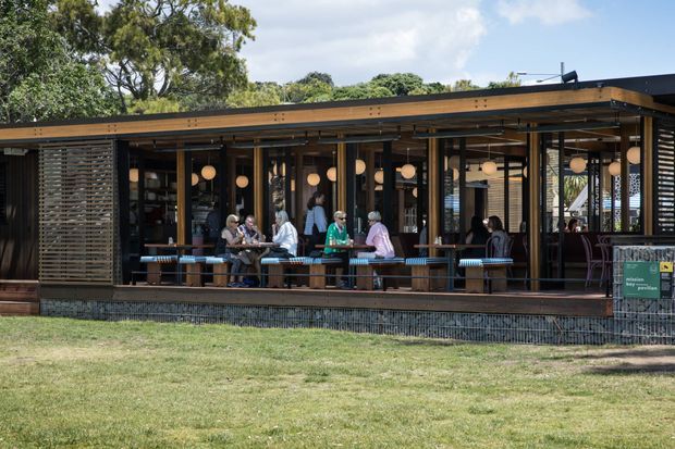 A low-slung contemporary pavilion-style café or hospitality building viewed from across a lawn. The structure features a broad flat roof with warm timber fascia, extensive floor-to-ceiling glazing, dark slender framing, timber decking, horizontal timber screening, and a long open dining counter or bar. Several people are seated inside beneath warm spherical pendant lights. Mature trees and lawn surround the building, while a small green sign is visible near the right entrance.