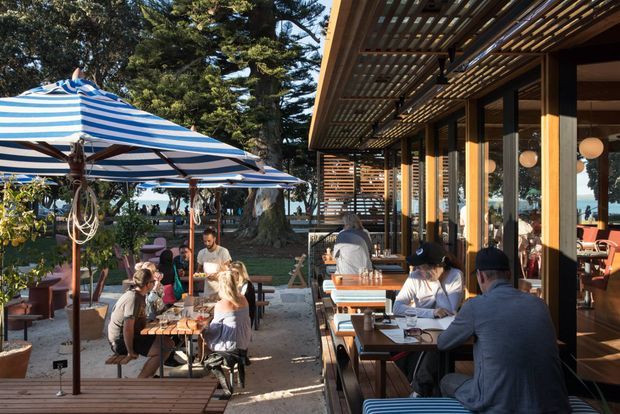 A lively outdoor dining terrace beside a glazed, timber-framed pavilion. Guests sit at timber tables beneath large blue-and-white striped umbrellas, with a deep timber pergola providing shade along the building facade. Coastal vegetation and glimpses of water are visible beyond the terrace.