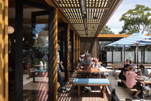 A lively outdoor dining terrace beside a glazed contemporary building. The space features timber decking, long communal timber tables and benches, a substantial timber pergola with a slatted overhead canopy, black-framed glazing, and striped blue-and-white umbrellas. Diners occupy the terrace, with an adjacent enclosed dining area visible beyond.