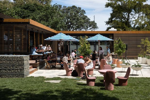 A lively outdoor dining and lounge courtyard beside a low, timber-clad contemporary building. The space features striped umbrellas, communal and individual seating, pink sculptural lounge chairs, potted greenery, a paved terrace, lawn, and a gabion-style stone retaining wall. Slatted timber screens and extensive glazing define the building perimeter, with mature trees forming a leafy backdrop.