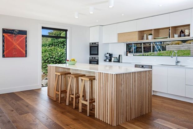 Bright contemporary kitchen with a large white island featuring vertical timber slat cladding and integrated seating for four. The room has white flat-panel cabinetry, a long white benchtop, a mirrored or glazed splashback reflecting greenery, timber flooring, recessed and pendant-style ceiling lighting, and a garden view through the doorway. Warm timber accents contrast with the predominantly white fit-out.