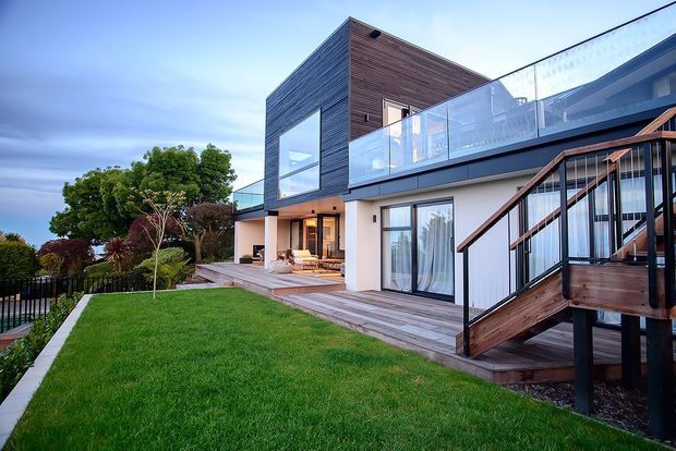 Exterior view of a contemporary multi-level home with a bold cubic upper volume clad in dark horizontal timber, white rendered walls, extensive glazing, and a glass-balustraded upper terrace. Timber decking and a timber stair connect the elevated terrace to a manicured lawn and landscaped garden.