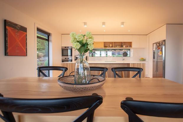 Warm, contemporary open-plan kitchen and dining area viewed from behind a timber dining table. The room features light timber cabinetry and flooring, a long kitchen island with integrated seating, stainless-steel appliances, a pale benchtop, recessed ceiling lighting, and large windows bringing in natural light. Black dining chairs and a glass vase provide contrast.