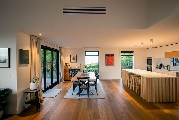 Open-plan contemporary kitchen and dining area with warm timber flooring, a large island with vertical timber-panelled cabinetry and bar seating, a dining table for six, full-height glazed doors opening to greenery, and light neutral walls. Artwork, plants, and simple pendant-free recessed lighting create a warm, refined interior.
