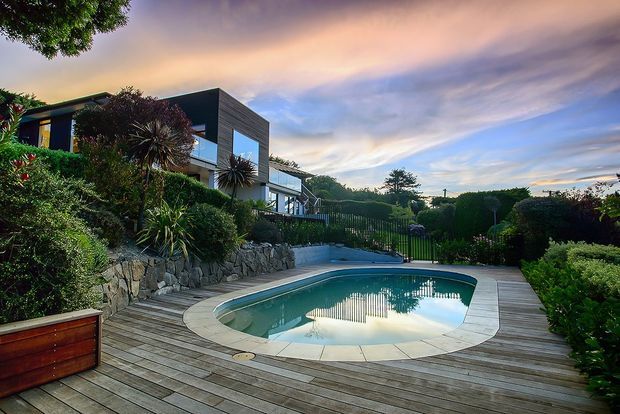 A modern multi-level home set in a landscaped, sloping garden, viewed from a timber pool deck. The image features a curved in-ground swimming pool, extensive planting, stone retaining walls, privacy hedges, and a dark-clad house with large glazed openings beneath a dramatic evening sky.