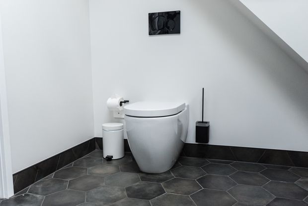A compact modern toilet room beneath a sloped ceiling, with a sculptural white toilet, white walls, dark hexagonal floor tiles, matching dark wall skirting, and minimal black bathroom accessories.