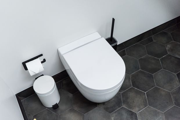 A compact, contemporary bathroom focused on a wall-mounted white toilet with matching white lid and adjacent small waste bin. Dark hexagonal floor tiles, white walls, and black toilet-roll and brush fittings create a clean, high-contrast scheme.