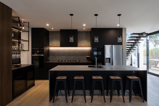A sophisticated contemporary kitchen with a large dark island and five bar stools, matte black cabinetry, integrated stainless-steel appliances, and a textured dark splashback with warm under-cabinet lighting. Open shelving and a built-in drinks cabinet occupy the left side, while a floating staircase and extensive glazing connect the kitchen to a bright adjoining outdoor area.