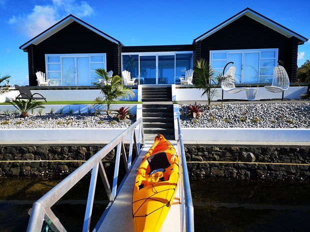 A symmetrical waterfront holiday-style residence viewed from a narrow dock, with a bright orange kayak in the foreground. The single-storey building has two dark-clad gabled wings connected by a glazed central section, broad white-framed windows, outdoor seating, white gravel landscaping, palms, and stepped access down to the water. The scene is sunny, crisp, and strongly coastal or resort-like.