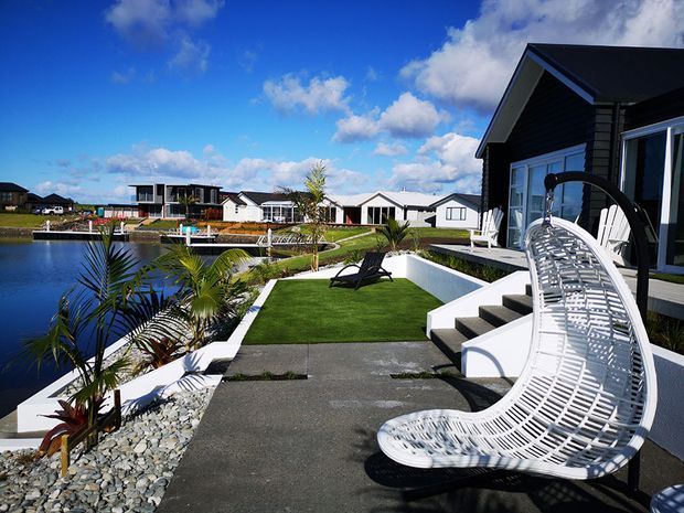 A modern dark-clad waterfront home with a landscaped outdoor terrace beside a narrow body of water. The space includes paved pathways, artificial lawn, tropical palms, white retaining walls, steps to the house, and a sculptural white outdoor lounge chair. Similar contemporary homes are visible across the water under a bright blue sky.