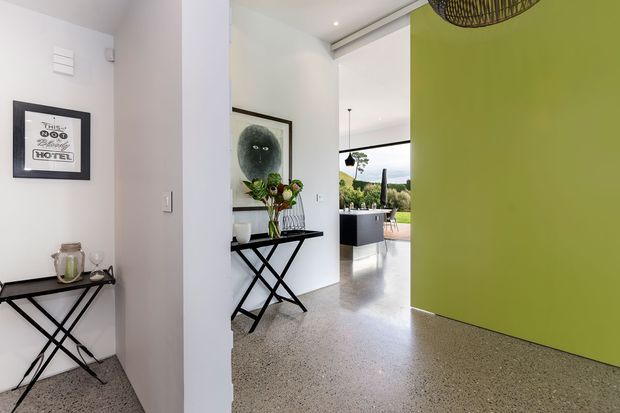 A bright contemporary interior entryway or hallway with polished terrazzo flooring, white walls, a vivid lime-green feature wall, and black metal console tables. The space opens toward a kitchen or living area with large glazing and greenery beyond. Framed artwork, decorative objects, and a woven pendant light add visual interest.