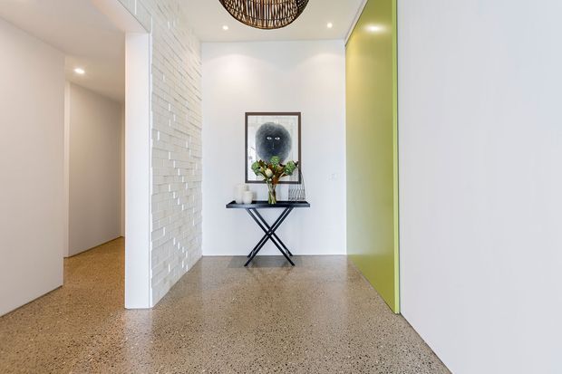 Bright contemporary entry hall with polished exposed-aggregate or terrazzo-style flooring, white walls, a white textured brick feature wall, and a vivid lime-green door or wall panel. A slim console table with artwork and decorative objects sits beneath a woven pendant light. An open hallway leads to another room on the left.