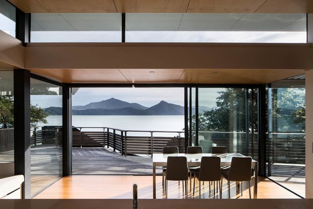 A contemporary open-plan dining area with a rectangular dining table and chairs, opening onto a timber deck through extensive floor-to-ceiling glazing. The room features warm timber ceilings and flooring, dark slender window and door frames, and a broad view across a lake toward mountainous terrain. A partially visible living area is at the left edge.
