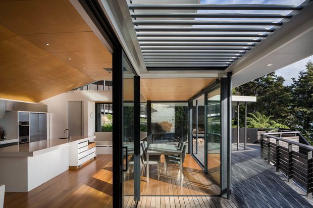A contemporary open-plan kitchen and dining area extending onto a timber deck through extensive black-framed glazing. The interior features a white island and cabinetry, warm timber flooring and ceiling lining, while an overhead glazed pergola creates a bright indoor-outdoor transition. The deck has dark horizontal privacy screens and overlooks landscaped trees.