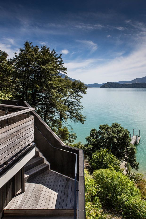 A multi-level timber deck and stairway descends through dense vegetation toward a calm turquoise lake, with forested hills and mountains across the water beneath a partly cloudy blue sky. The image is an exterior architectural landscape view dominated by the deck, balustrades, stairs, and lakeside setting.