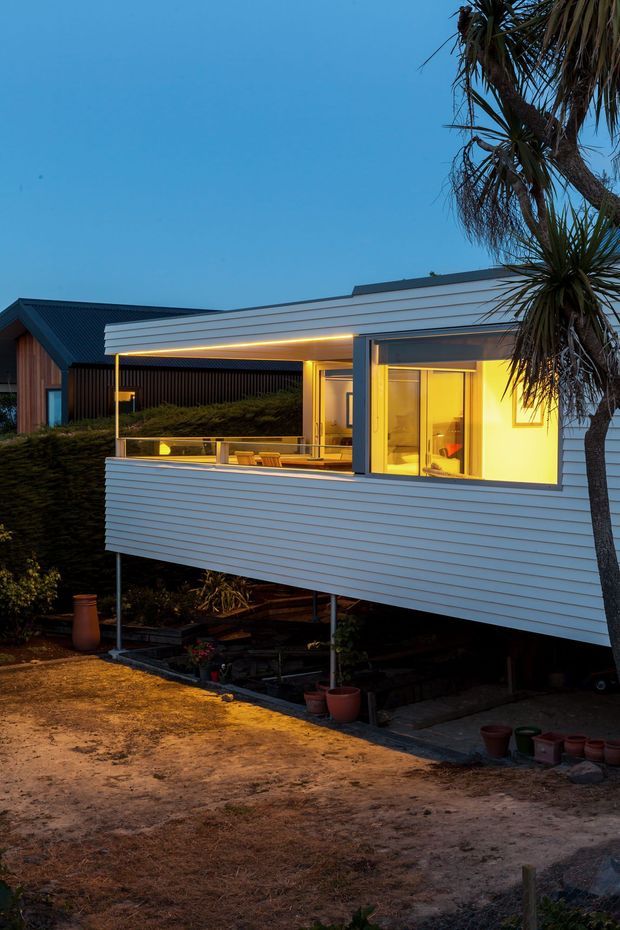 Twilight exterior view of a compact elevated modern home or extension with white horizontal weatherboard cladding, a covered balcony or terrace, expansive glazing, and warm interior lighting. The building is raised on slender supports above a landscaped sandy site, with a dark pitched-roof structure and coastal-style trees visible nearby.