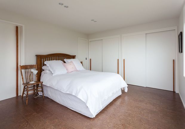 Bright, spacious bedroom with a white upholstered bed dressed in white bedding and pale pink accent pillows. The room has warm brown timber flooring, white walls and ceiling, built-in white wardrobes with long timber handles, a classic timber headboard, and a wooden chair beside the bed. Recessed ceiling lights and simple traditional detailing create a calm, understated interior.