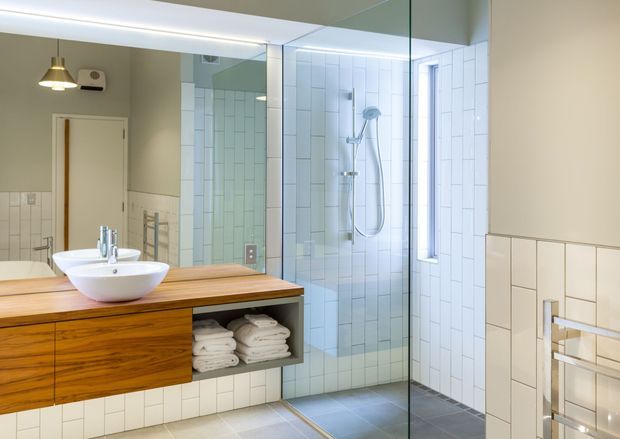 Bright contemporary bathroom with a floating timber vanity, two white vessel basins, open towel storage, a large frameless glass shower enclosure, white rectangular wall tiles, grey floor tiles, and integrated wall lighting. A wall-mounted towel rail and partially visible bathtub or ledge appear at right.