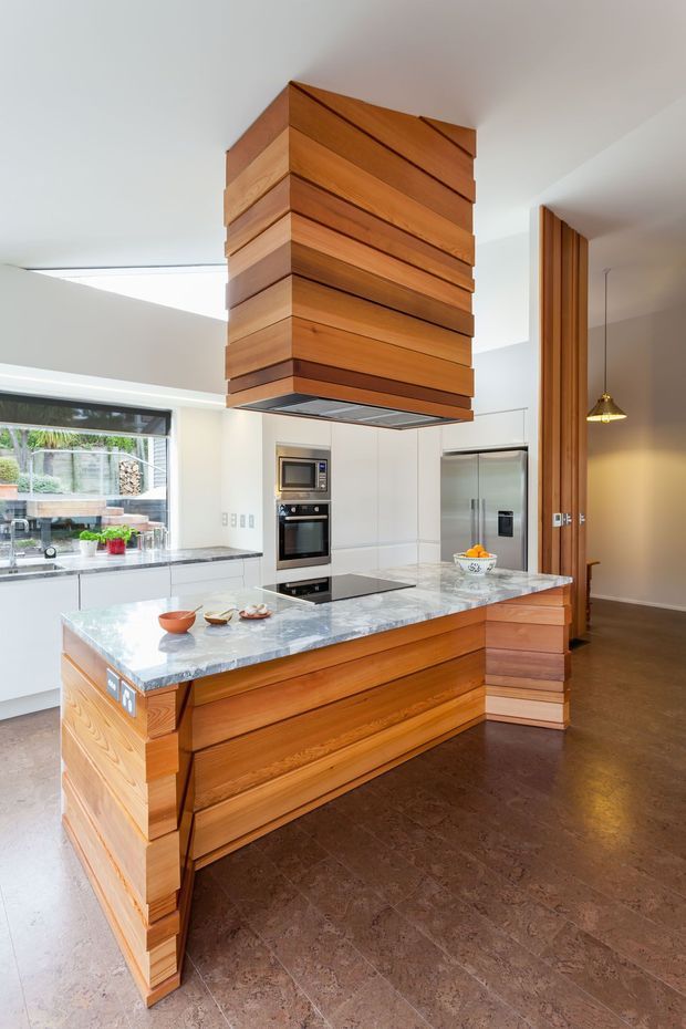 A bright contemporary kitchen dominated by a sculptural central island and matching rangehood enclosure clad in layered horizontal timber boards. The island has a pale veined stone benchtop, warm timber cabinetry, integrated appliances, stainless-steel refrigeration, dark stone-look flooring, and white walls. A glazed opening looks onto an outdoor area, while a timber-lined opening leads toward an adjacent space.