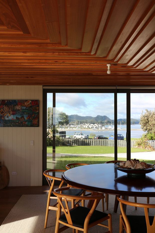 A warm, wood-lined dining room with a large round dark timber dining table and matching wooden chairs. The room has extensive timber ceiling detailing, vertical wall panelling, carpet flooring, and full-height black-framed sliding glazing opening to a lawn and waterfront marina view with hills beyond. A colourful artwork hangs on the left wall.