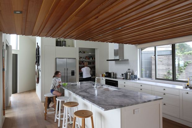 Bright contemporary kitchen with a large marble-look island and integrated breakfast seating, white built-in cabinetry, stainless-steel refrigerator, open shelving, and broad windows. A striking slatted timber ceiling adds warmth and texture, while pale timber flooring and abundant daylight create an inviting open-plan space.