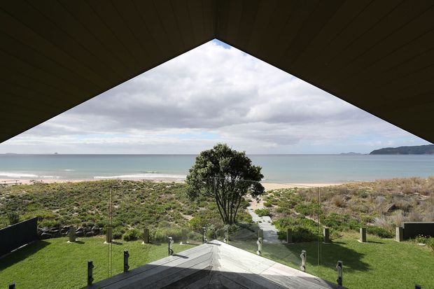 View from beneath a dark, dramatically angled roof overhang toward a landscaped coastal property, with a central paved path leading through lawns and dune planting to a sandy beach and calm sea. A distant headland is visible across the water under a cloudy sky.