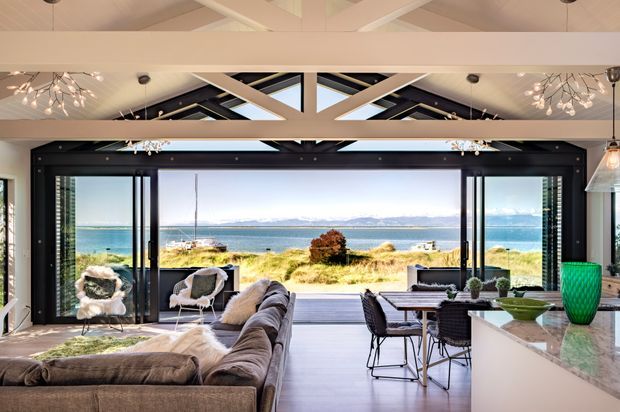 Open-plan coastal interior with a living room, dining area and kitchen arranged around expansive glazed doors overlooking a beach and water. The room features a large sectional sofa, dining table, marble-look island benchtop, dark-framed glazing, exposed white structural roof beams, skylights and statement pendant lighting.