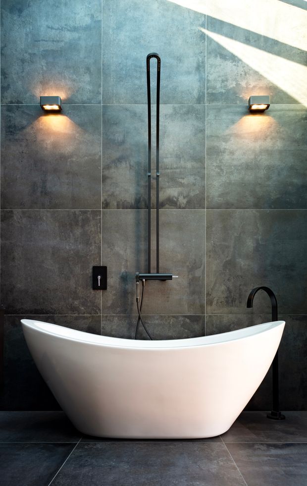 A dramatic contemporary bathroom featuring a sculptural freestanding white bathtub against large-format dark grey concrete-look wall tiles. A tall black wall-mounted shower fitting and separate black floor-mounted bath tap provide strong vertical accents, while two warm wall sconces illuminate the textured wall surface. Dark grey floor tiles and directional daylight create a moody, luxurious atmosphere.