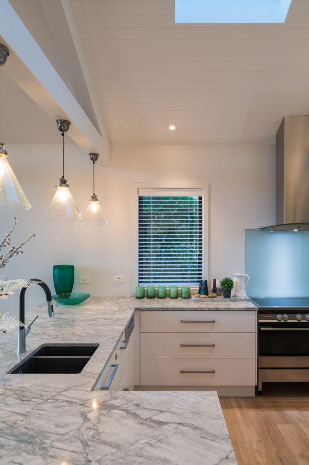 A bright, contemporary kitchen featuring extensive white-and-grey veined stone benchtops, pale built-in cabinetry, a black undermount sink with gooseneck tap, stainless cooking appliances, timber flooring, pendant lights, and a skylight. White plantation blinds cover the window, with green glassware and small decorative items adding colour.