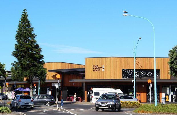 Street-level exterior view of a contemporary single-storey commercial building with warm vertical timber cladding, deep dark roof overhangs, and a prominent sign reading “The Rabbit Hole.” The building fronts a busy parking and street area with vehicles, pedestrian access, streetlights, landscaping, and a large evergreen tree.