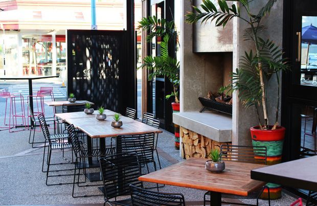 An urban outdoor dining area with multiple timber-topped tables and black wire-frame chairs arranged beside a storefront. Large potted tropical plants, a textured concrete feature wall, a built-in fireplace or planter-like niche with stacked firewood, and black-framed glazing create a contemporary café or hospitality setting. The scene is bright with a lively, casual atmosphere.