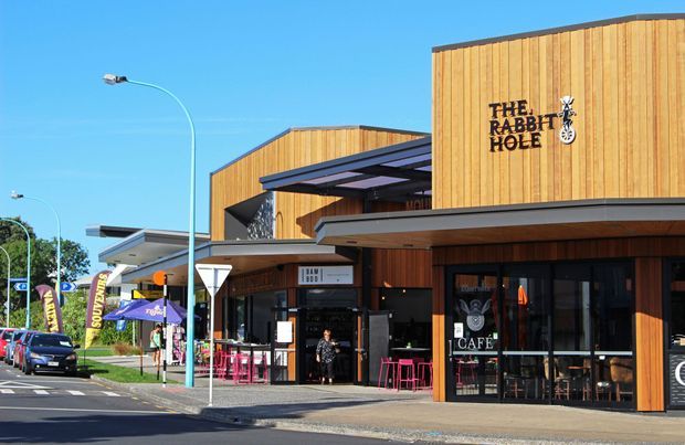 Street-level exterior view of a contemporary timber-clad commercial building containing The Rabbit Hole café and other retail or hospitality spaces. The building has strong horizontal roof canopies, dark metal framing, glazed shopfronts, and a layered, low-rise composition along an urban street.