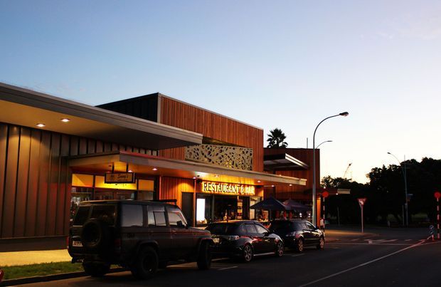 Street-level dusk photograph of a contemporary commercial restaurant building, with dark metal cladding, warm timber-toned wall panels, a patterned decorative screen, illuminated restaurant signage, covered entry canopy, outdoor umbrellas, parked vehicles, and a suburban street setting.